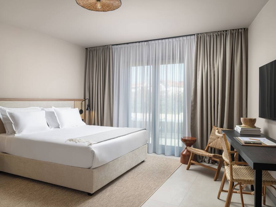 A hotel room with a large bed, a desk, wicker chairs, and a view through sheer curtains.