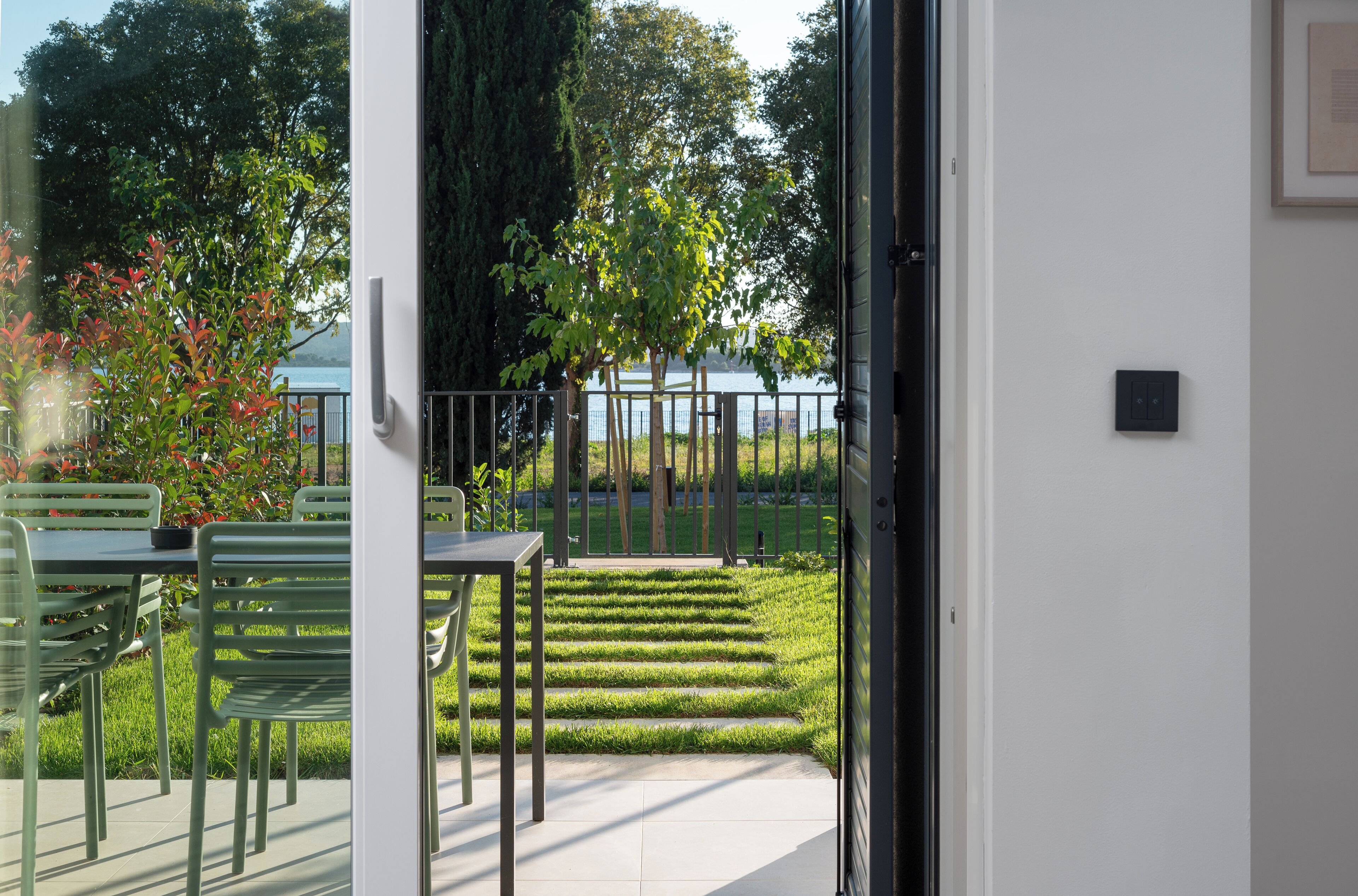 Eine Terrasse mit Tisch und Stühlen, ein kleiner Garten mit Baum und Blick auf das Meer durch eine Glasdoor.