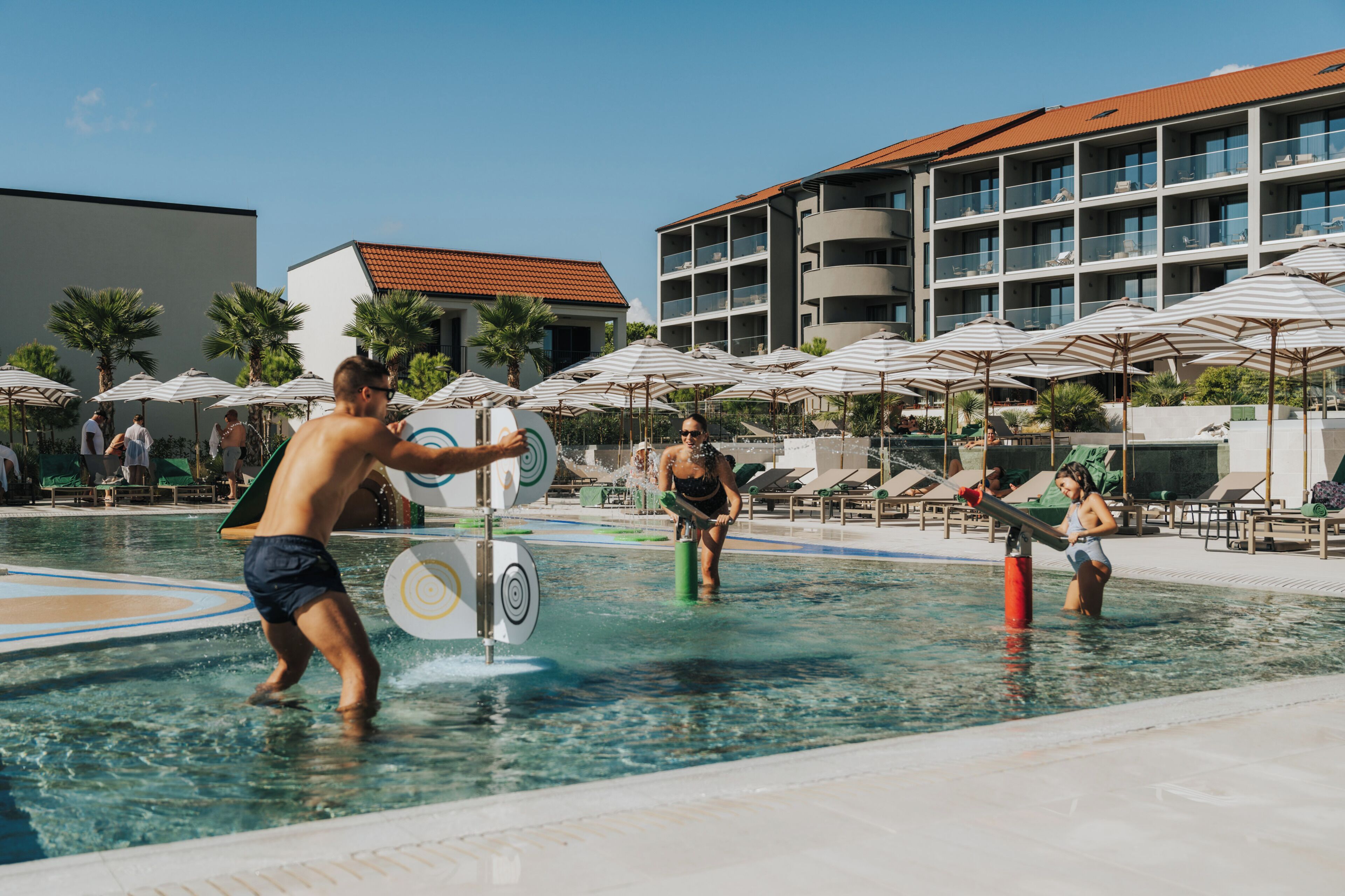A swimming pool area with lounge chairs and umbrellas, a building with balconies, and a clear sky.