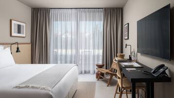 Quarto de hotel com uma cama grande, uma escrivaninha com telefone, uma cadeira e uma janela ampla com cortinas.