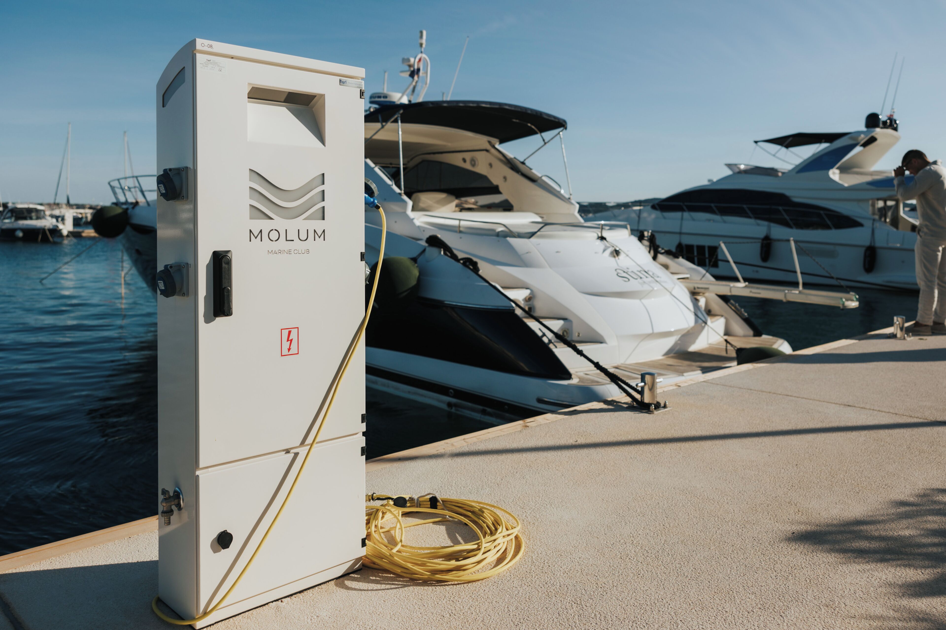 A marina with yachts docked, a white electrical box labeled 'MOLUM MARINE CLUB,' and a person taking a photo.