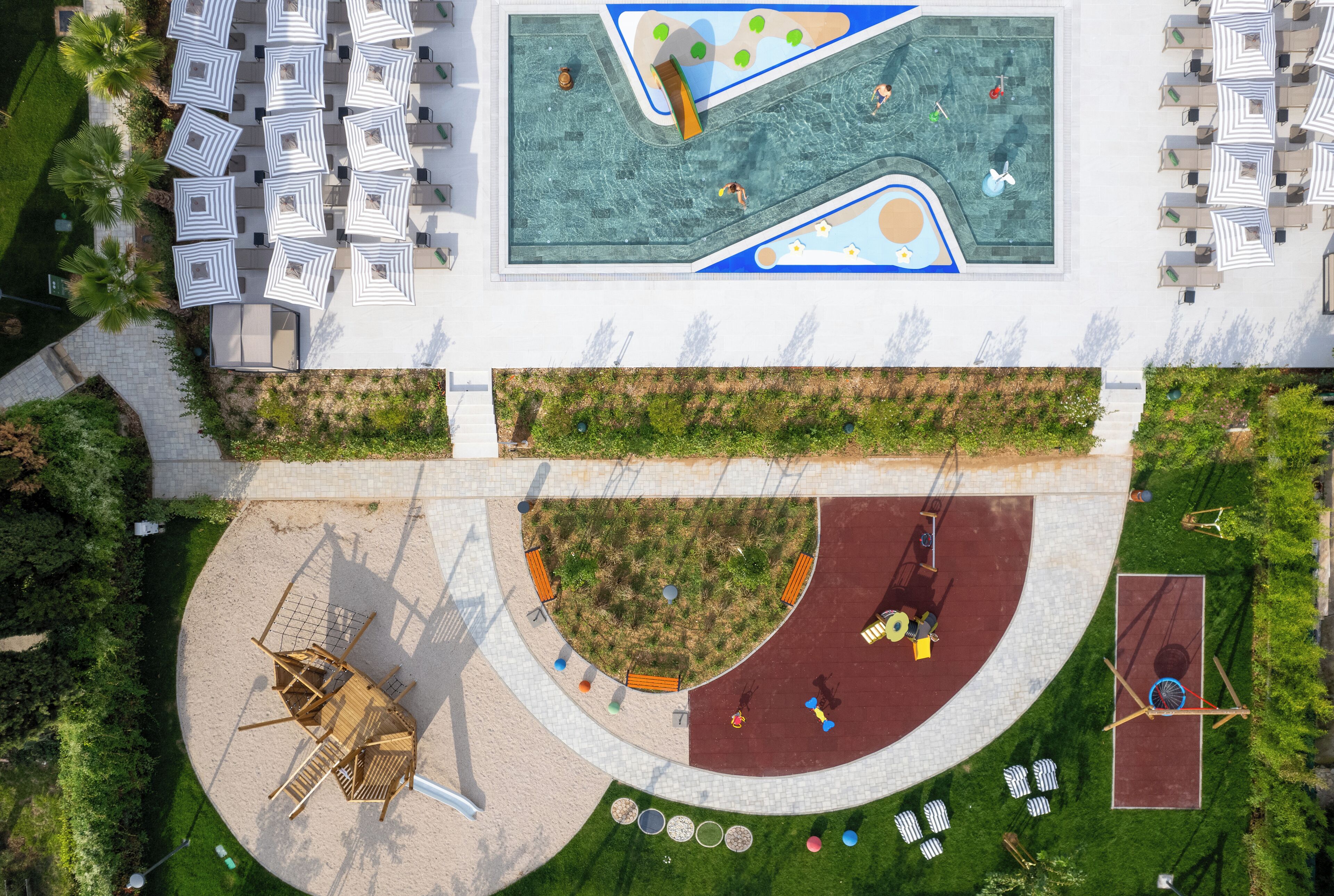 Vue aérienne d’un hôtel doté d’une piscine, de chaises longues, d’une aire de jeux et d’un jardin aménagé.