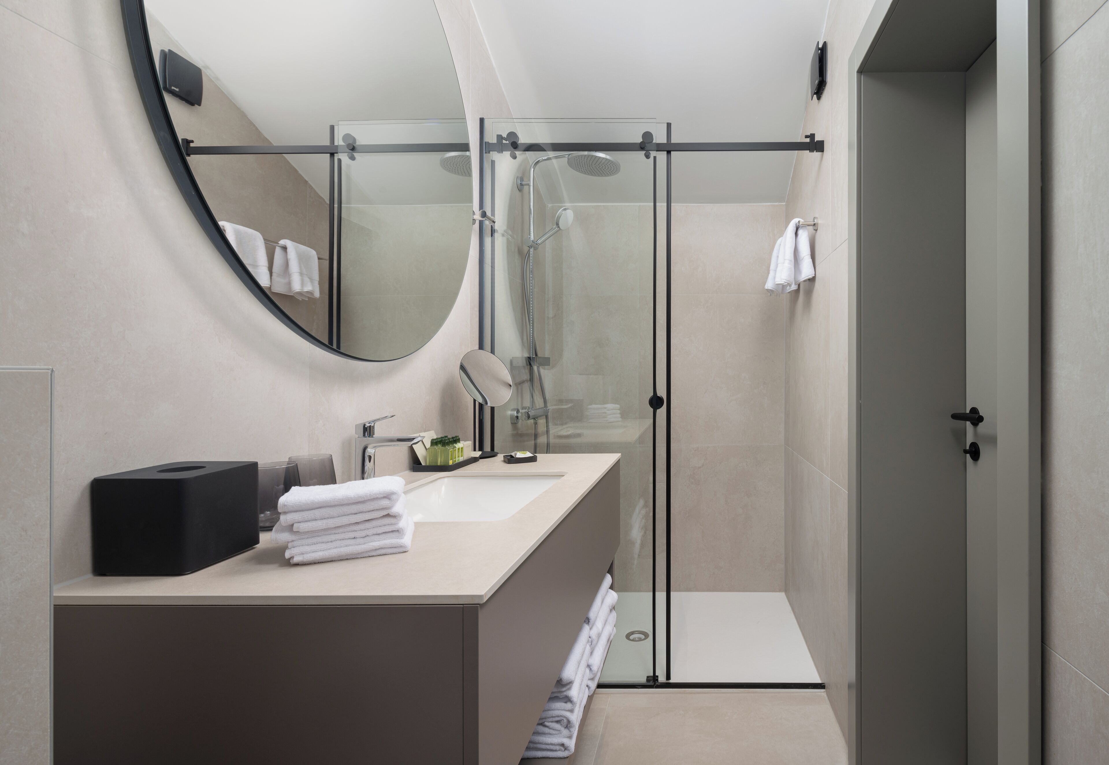 Une salle de bain moderne avec une douche à parois de verre, un miroir rond et un lavabo agrémenté de serviettes soigneusement pliées.
