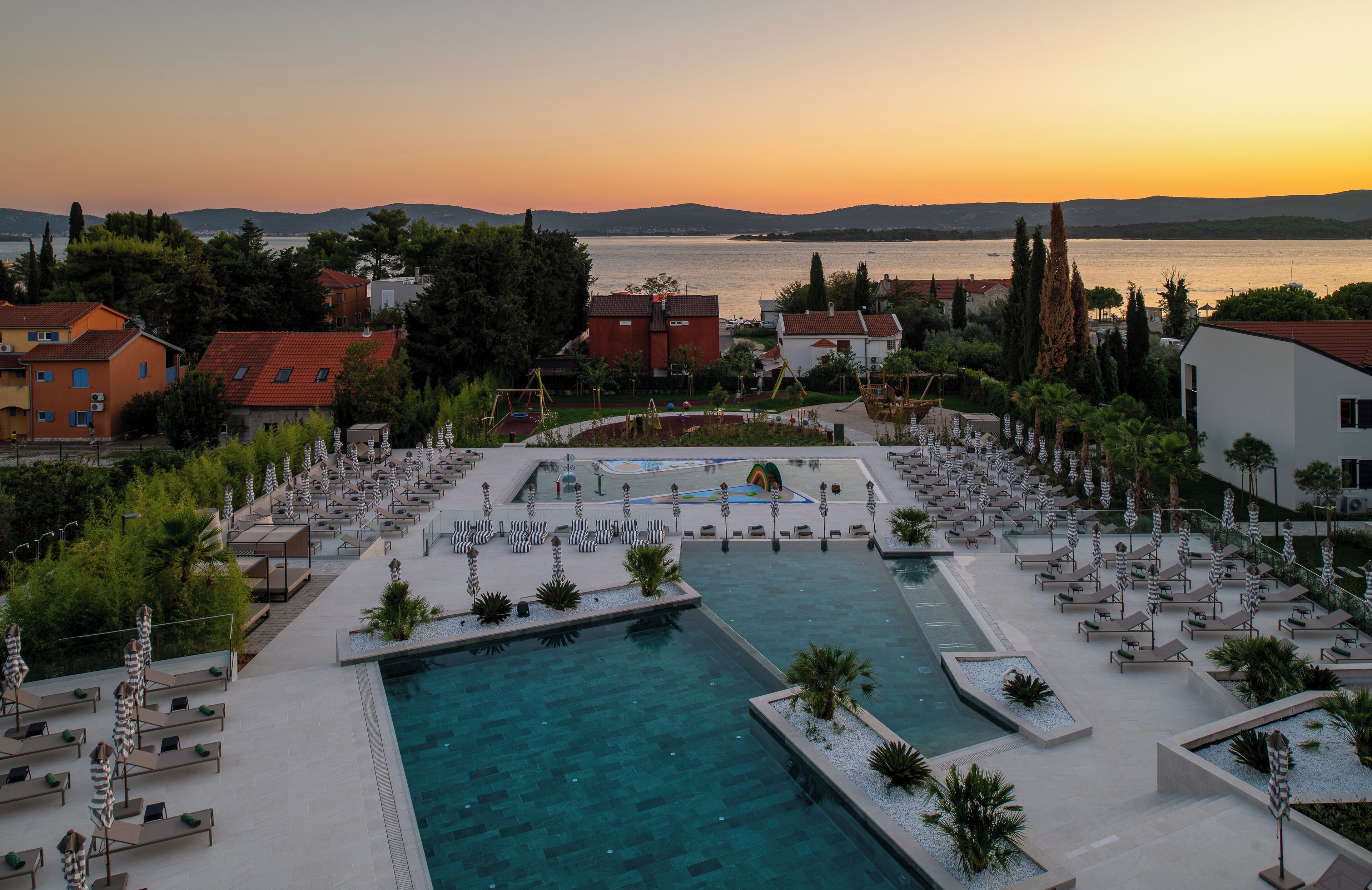 A resort with a swimming pool, lounge areas, and a view of the sea at sunset.