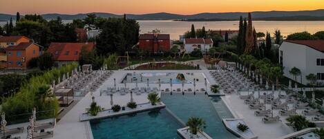 A resort with a swimming pool, lounge areas, and a view of the sea at sunset.