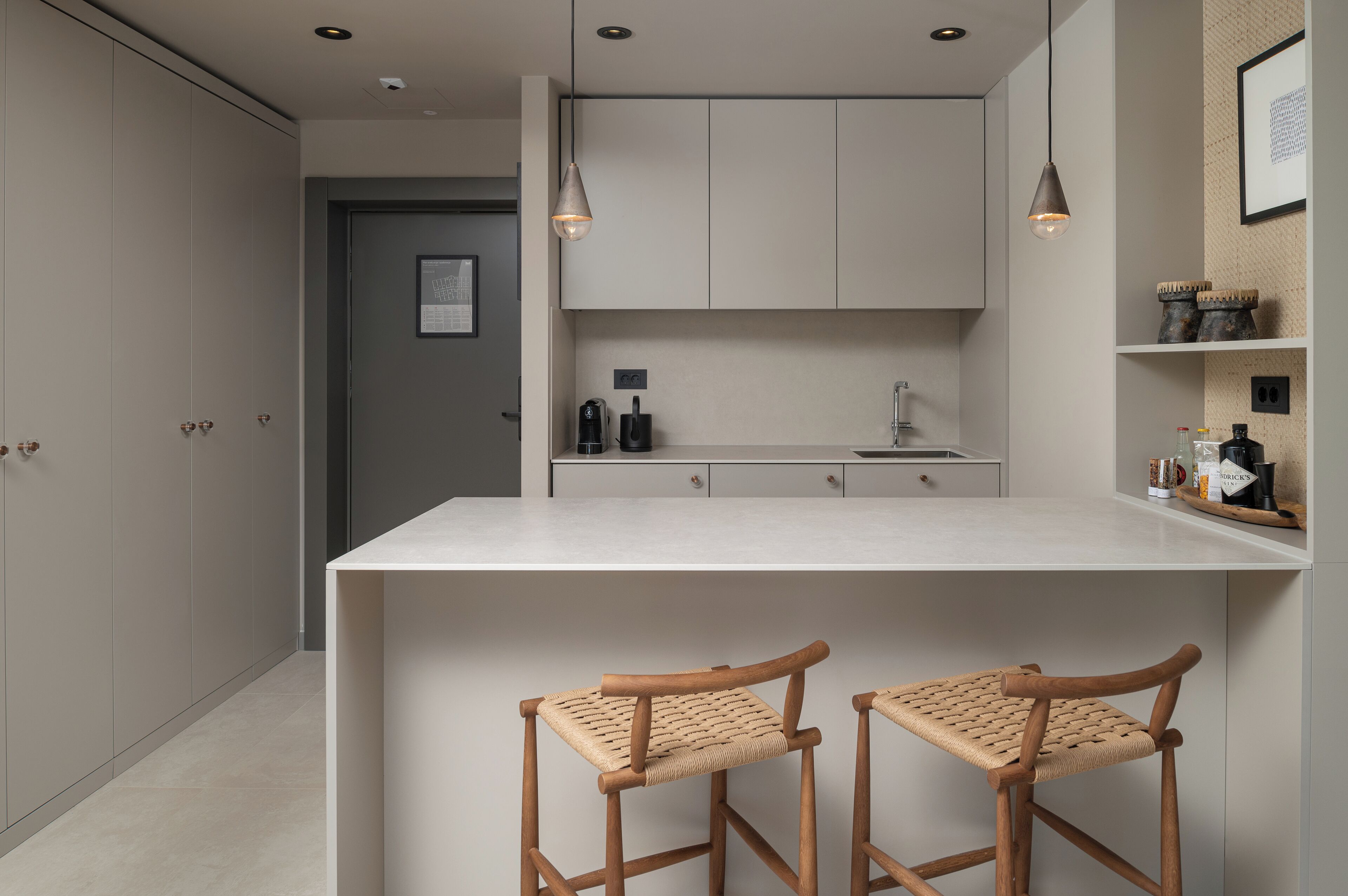 Una cocina moderna con una isla central, dos taburetes de madera, iluminación colgante y vistas al cuarto contiguo.
