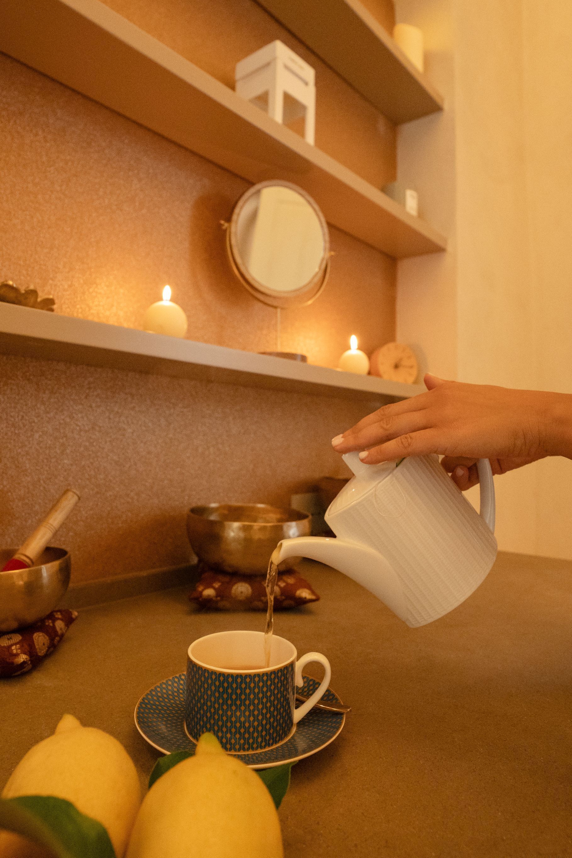 A hand pouring tea into a cup on a table with a candle, a bowl, and lemons.