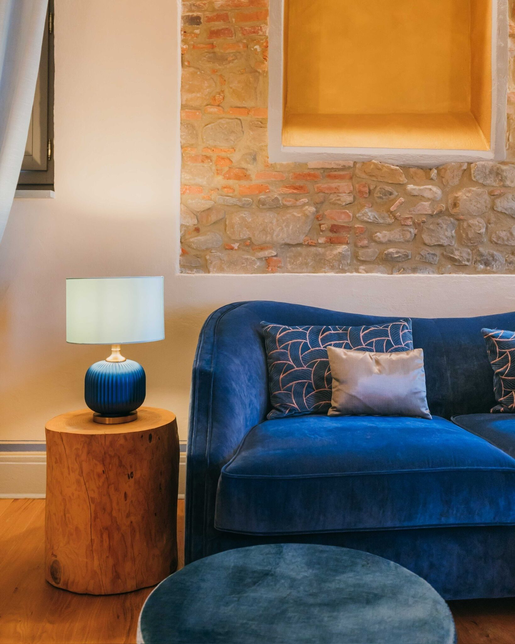 A room with a blue velvet sofa, a round wooden side table with a blue lamp, and a stone wall.