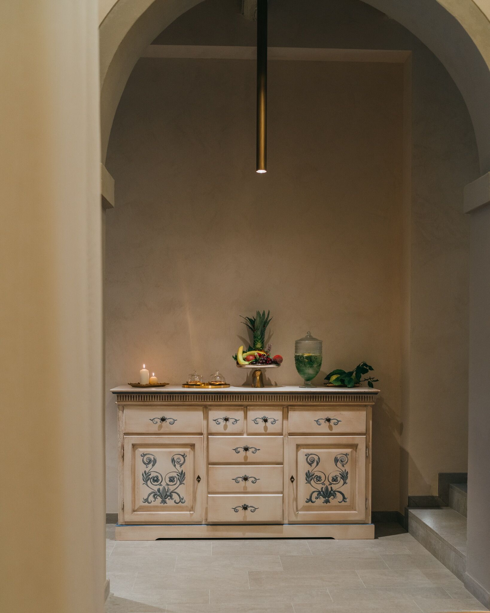 A hallway with a cream-colored cabinet featuring ornate blue designs, a hanging pendant light, and a fruit display on top.