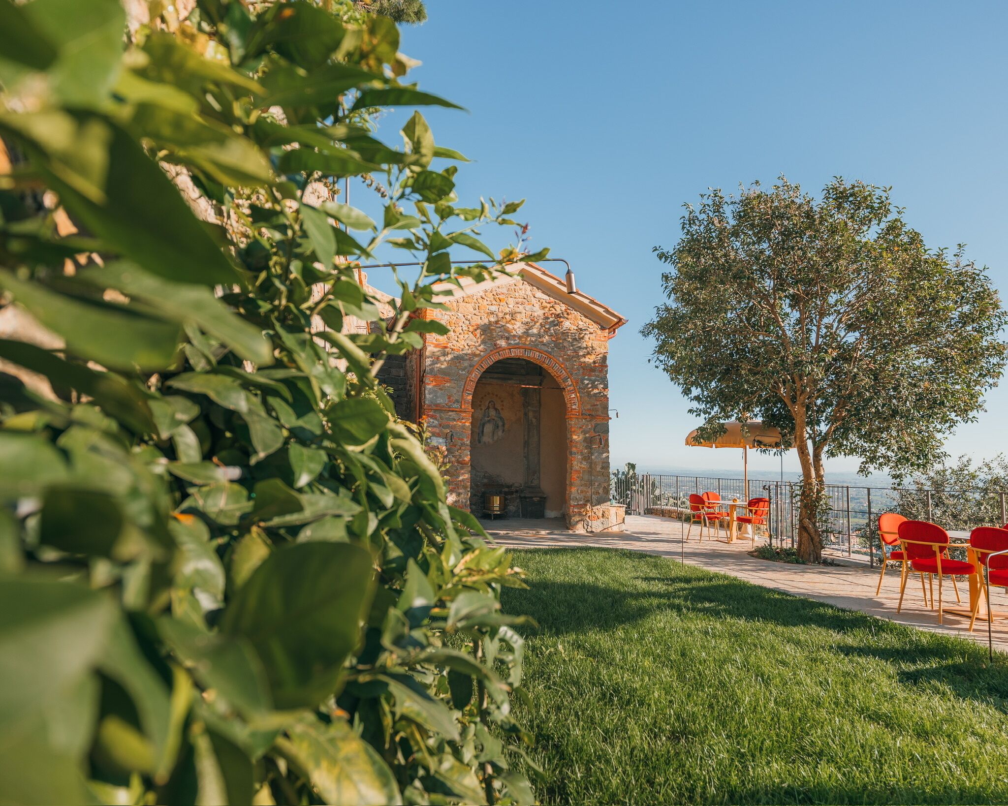 Un bâtiment en pierre avec une entrée en arc de cercle, une pelouse et des chaises rouges sous un arbre.