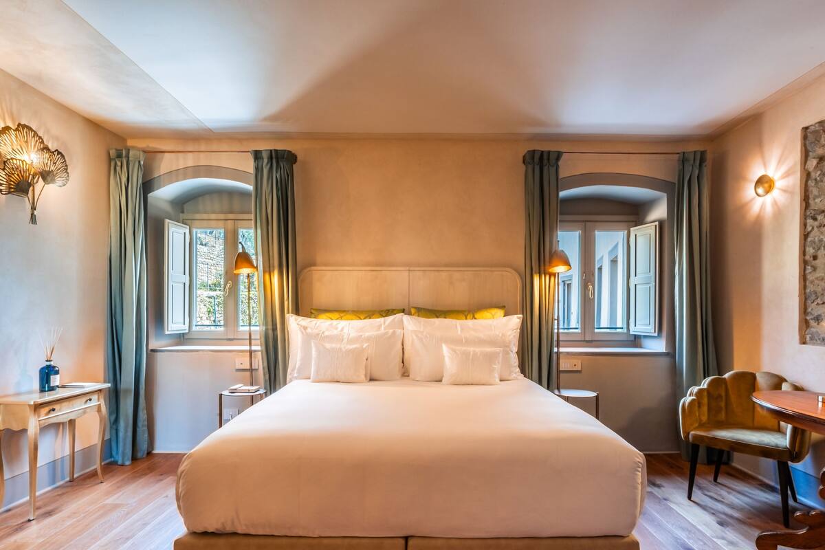 A large bed with white linens and multiple pillows, flanked by two windows with curtains, a bedside table, and a chair.