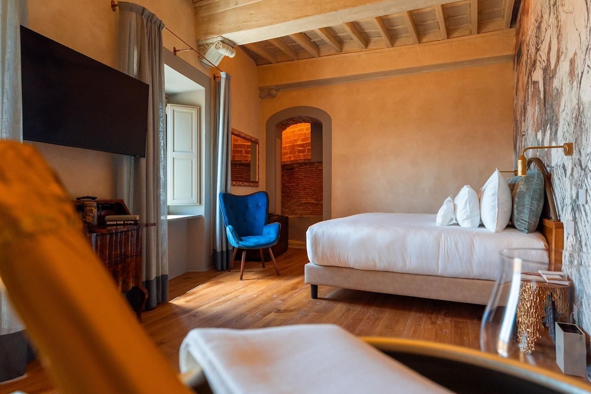 A cozy hotel room with a bed, a blue armchair, a wooden floor, and a brick wall.