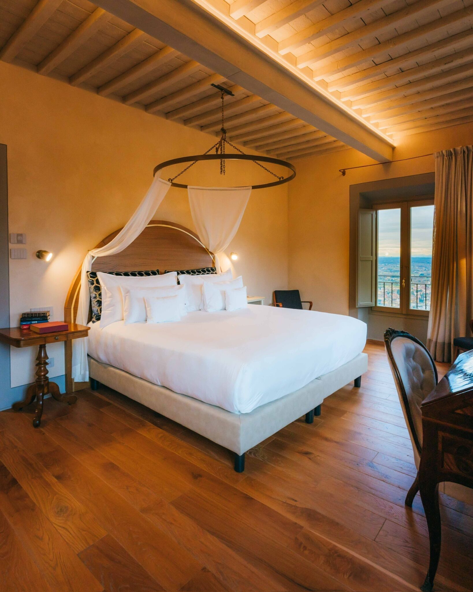 Un dormitorio amplio con una cama grande, piso de madera, dosel sobre la cama, una mesita de noche, una silla y una ventana con vistas.