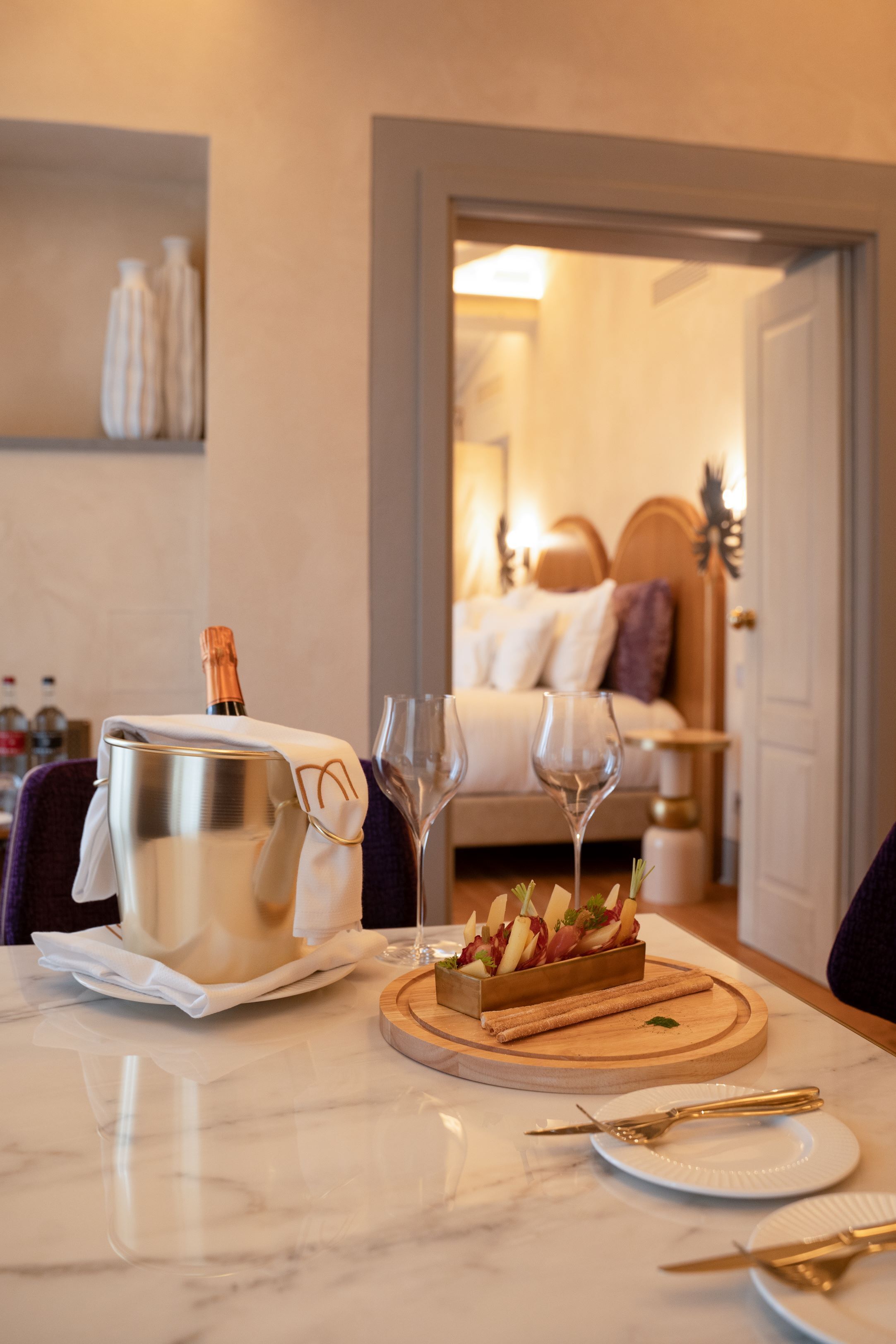 Ein Marmortisch mit einem Champagnerkühler, zwei Weingläsern, einer Holzplatte mit Vorspeisen und einem Bett im Hintergrund.