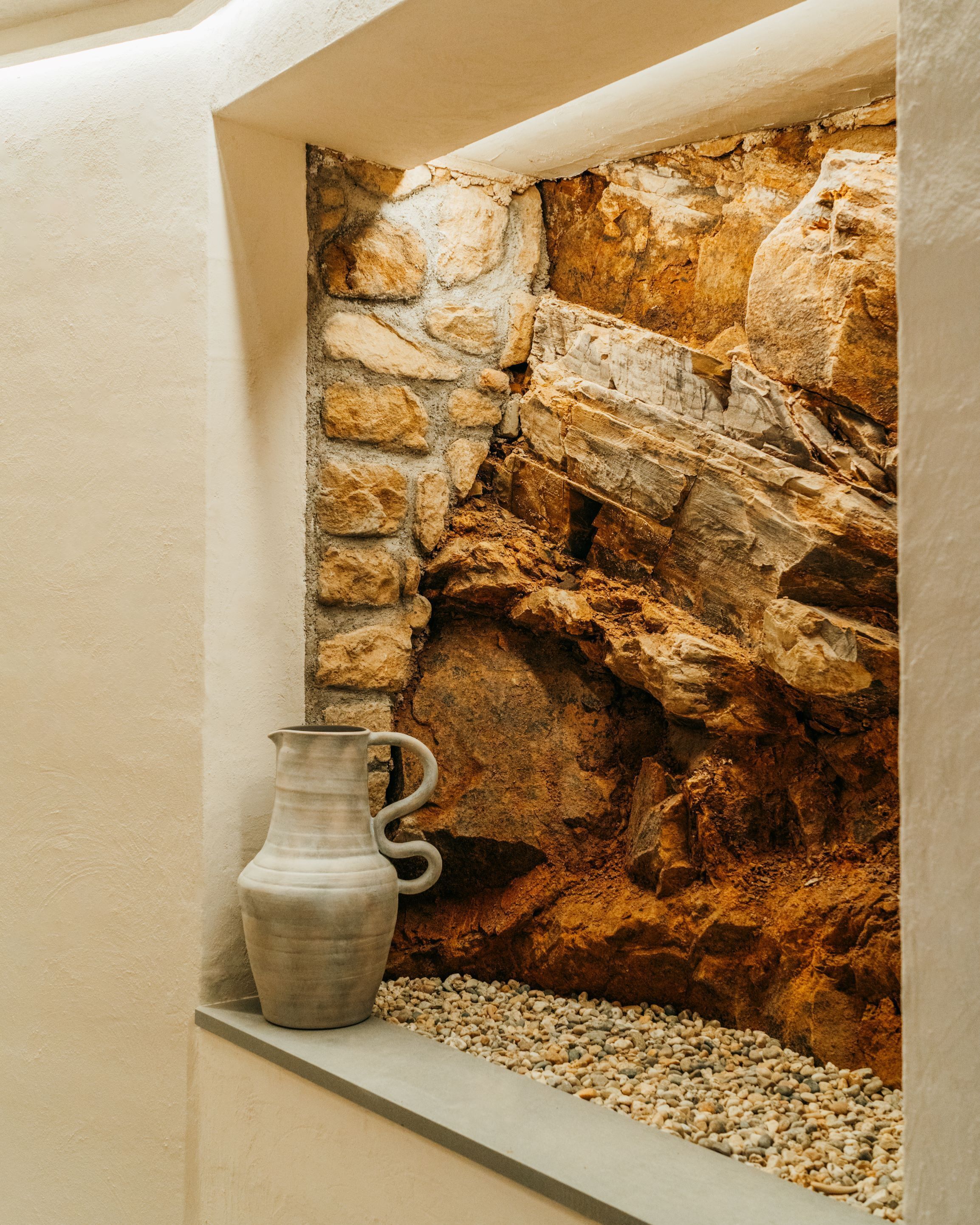 Un muro de piedra con aspecto rústico y texturizado, un pequeño estante con una jarra de cerámica y una zona con guijarros abajo.