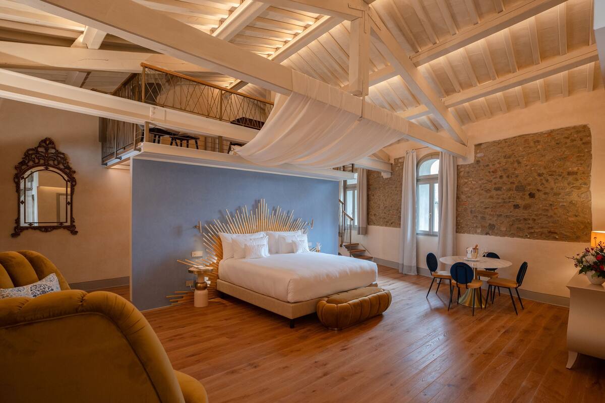 A spacious bedroom with a large bed, a dining area with chairs, a staircase, and a stone wall.