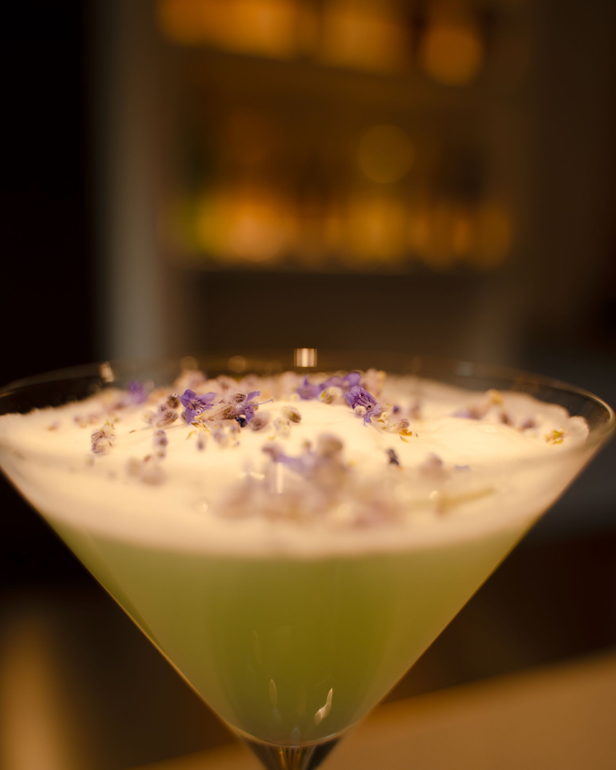 Un cóctel en una copa de martini con líquido verde y espuma blanca, adornado con flores púrpuras.