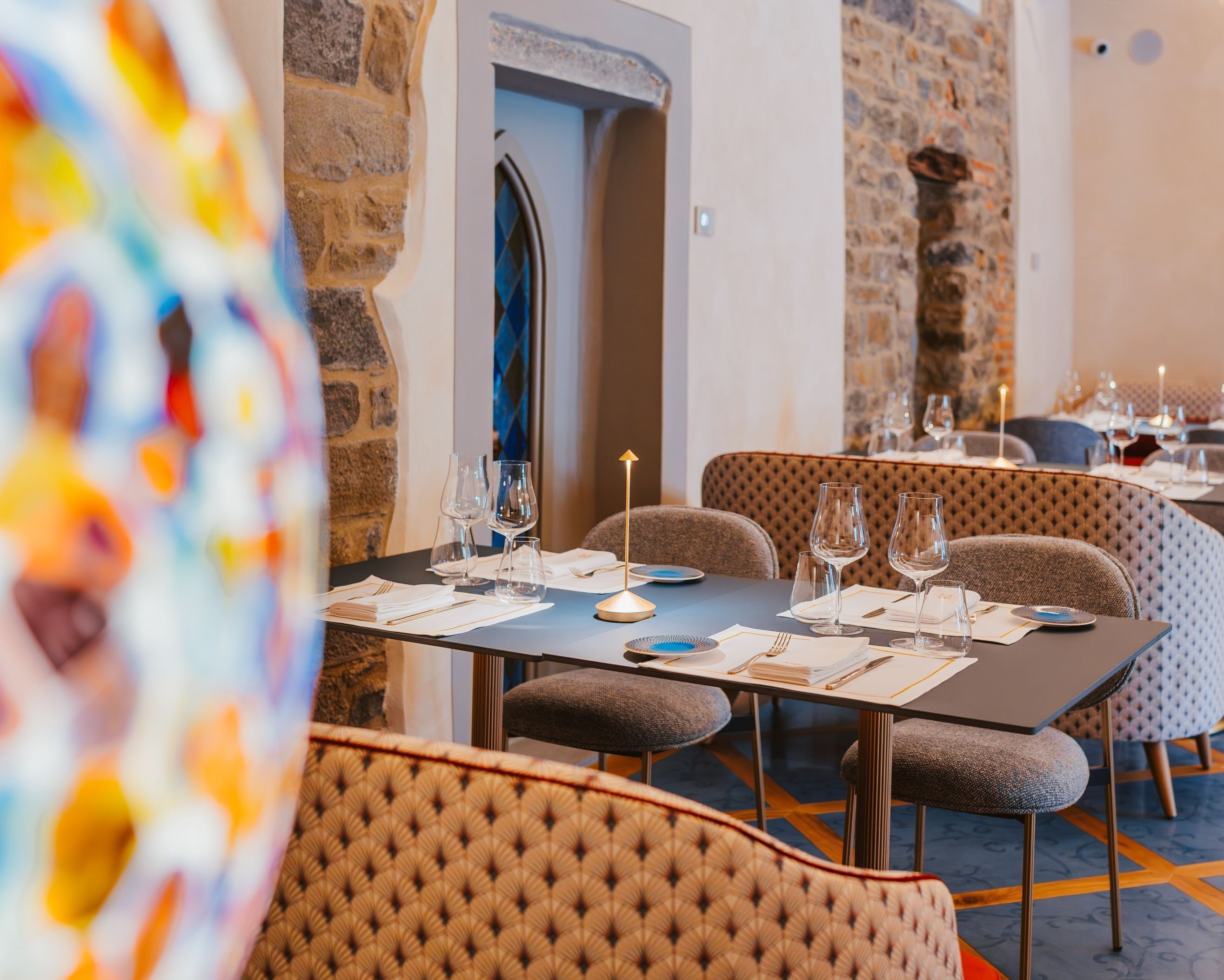 Ein Restaurant mit einer Steinwand und einem blau gemusterten Fenster. Tische sind mit Weingläsern, Tellern und Besteck gedeckt.