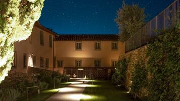 A courtyard with a pathway leading to a building, surrounded by greenery and illuminated by night lighting.