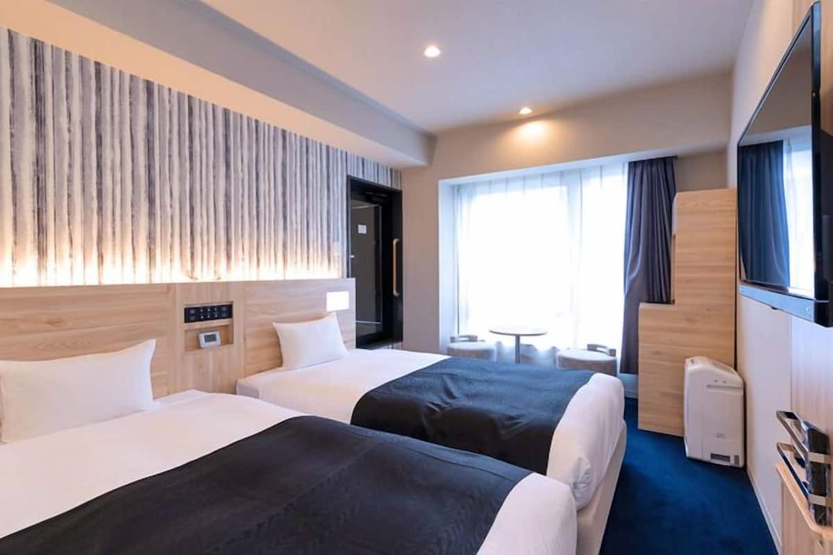 Quarto de hotel com duas camas, TV, máquina de café e um pequeno espaço de estar com mesa e cadeiras.