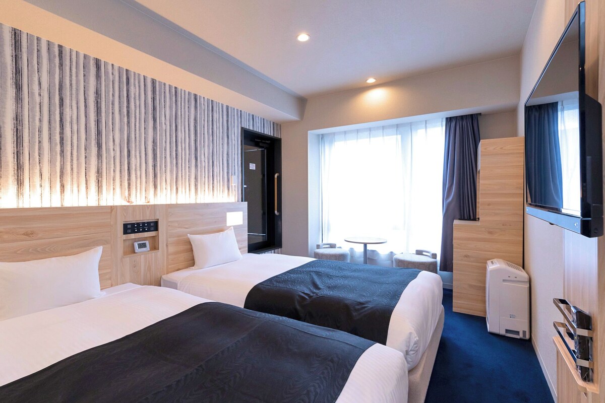 Habitación de hotel con dos camas, televisión, escritorio y ventana con cortinas.