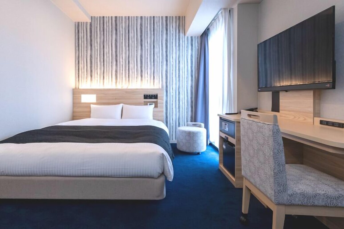 Habitación de hotel con cama, escritorio, silla, televisor y ventana con cortinas.