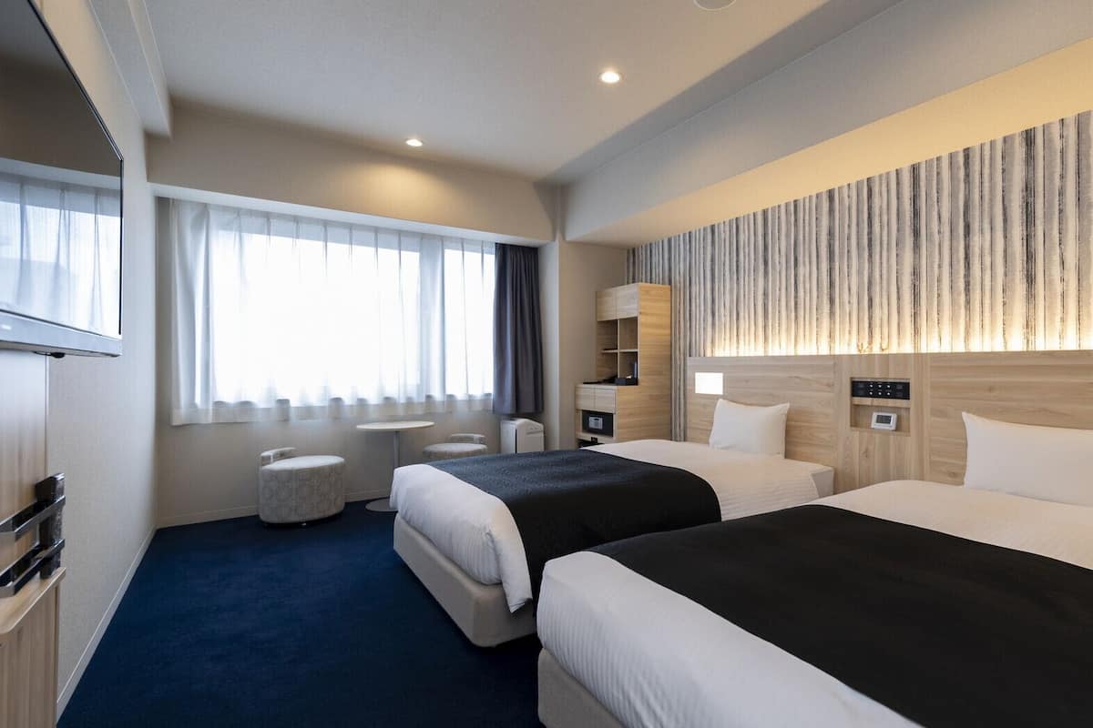 Quarto de hotel com duas camas, televisão, escrivaninha e janela com cortinas.