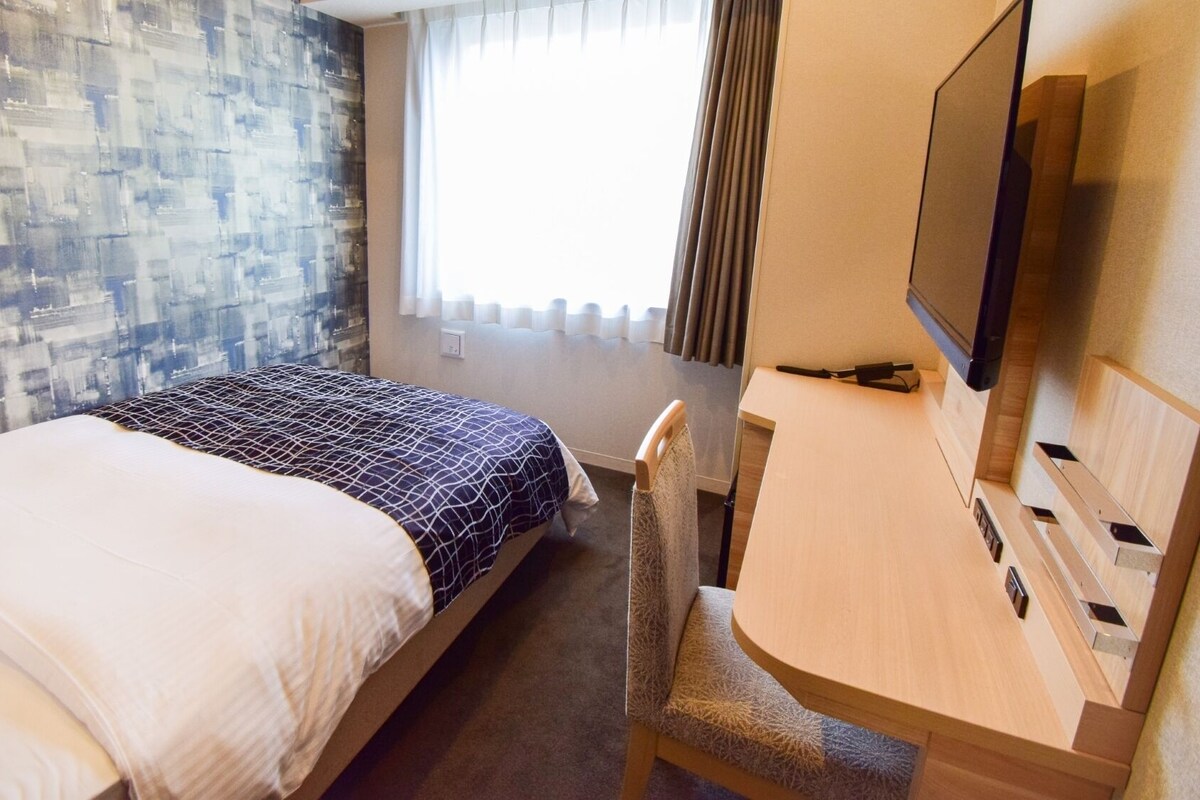 Habitación de hotel con cama, escritorio, silla, televisión y ventana con cortinas.