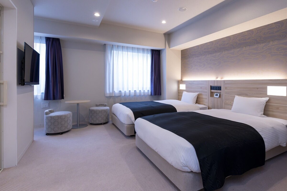 Habitación de hotel con dos camas, televisión, una mesita y ventana con cortinas.