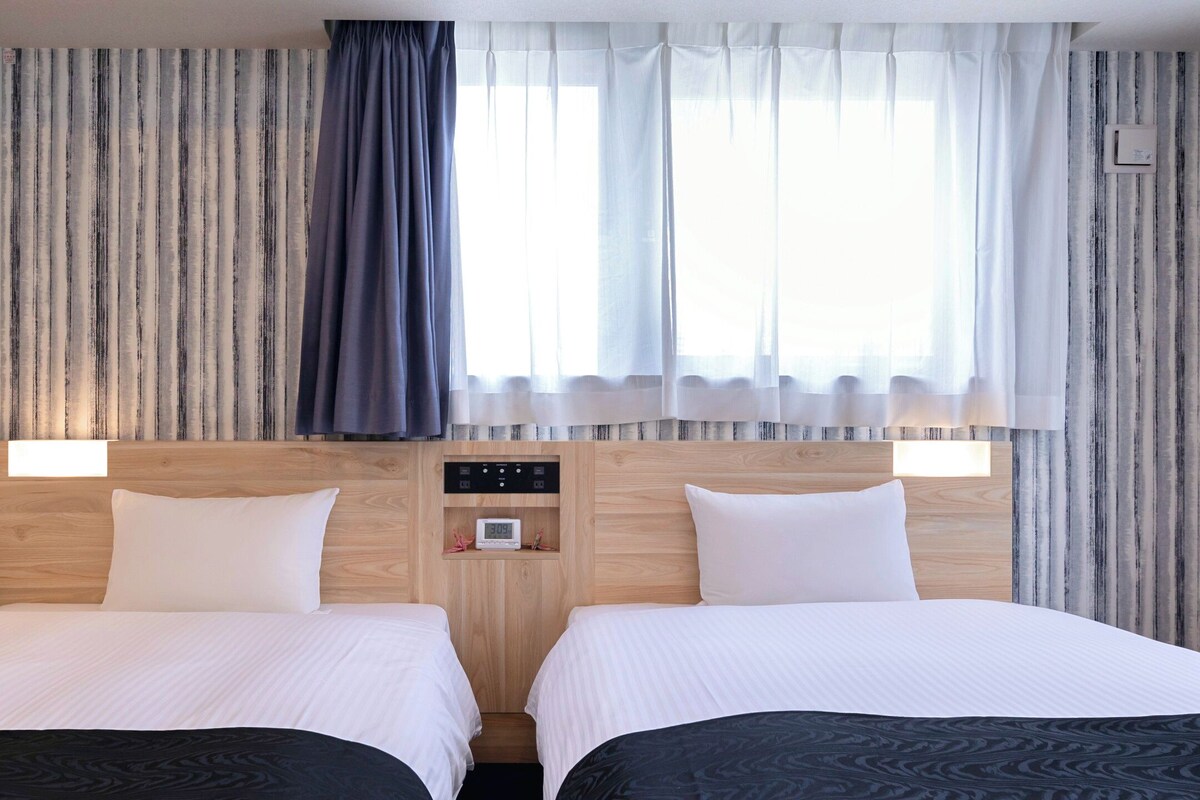 Habitación de hotel con dos camas, cabecero de madera, reloj digital y cortinas a rayas.
