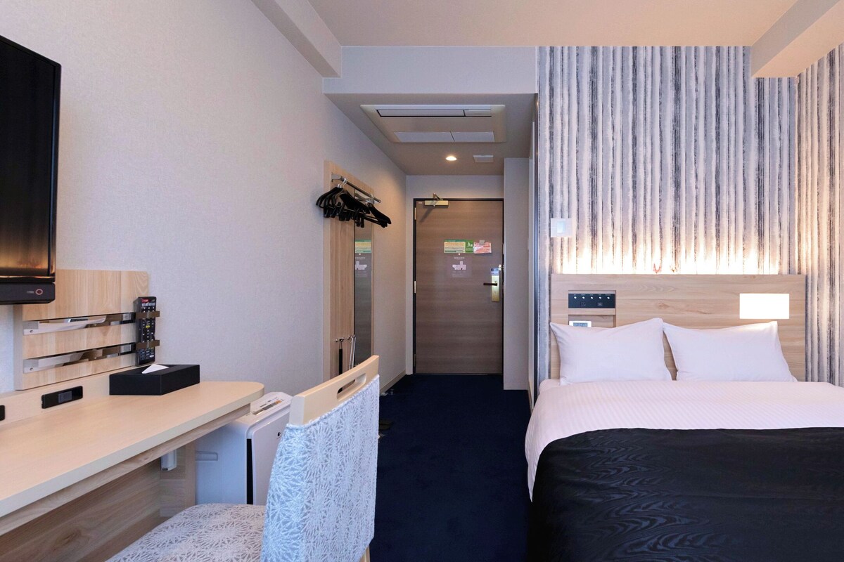 Habitación de hotel con cama, mesita de noche, televisor y vistas al pasillo.