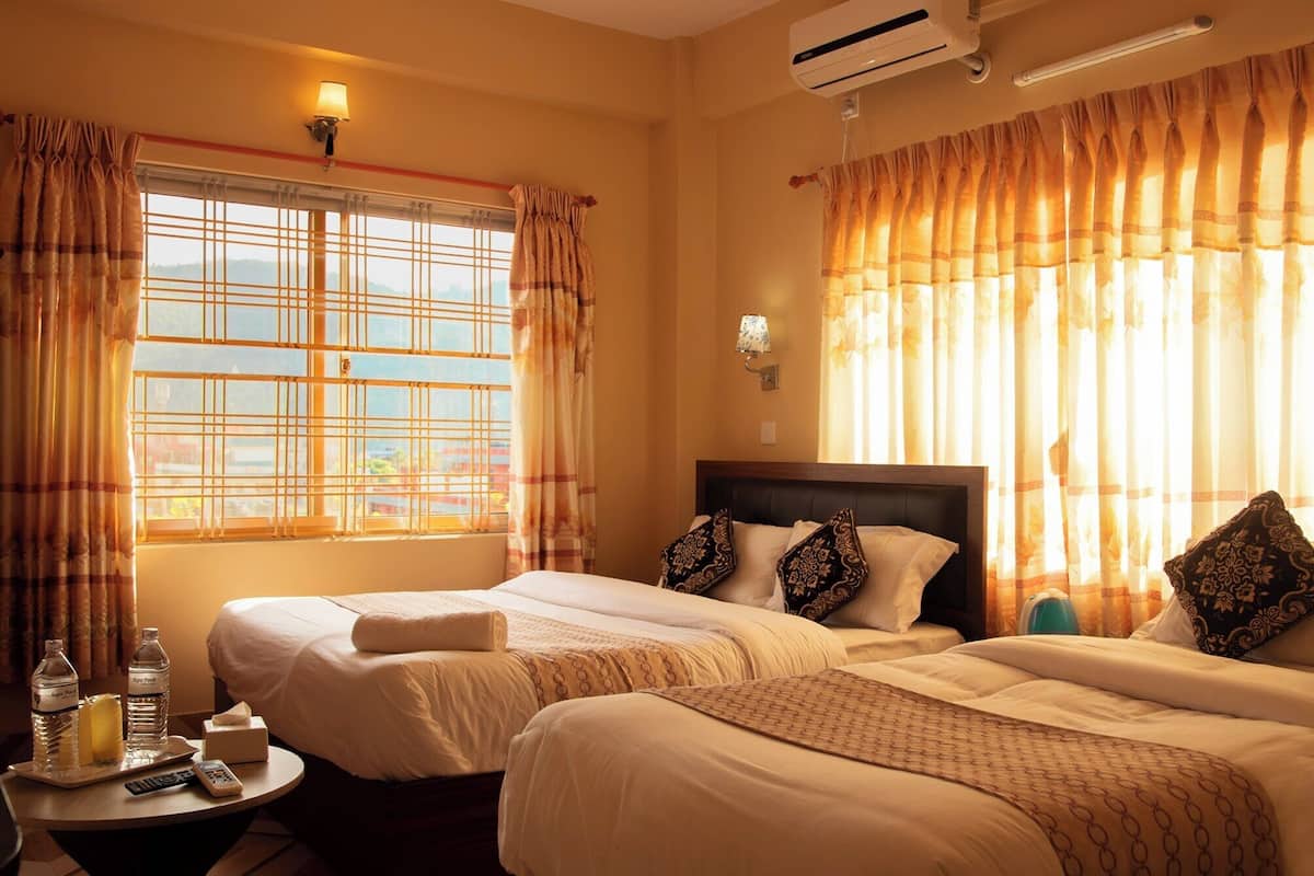 A hotel room with two beds, a bedside table with a bottle of water and a remote control, and a wall-mounted air conditioner.