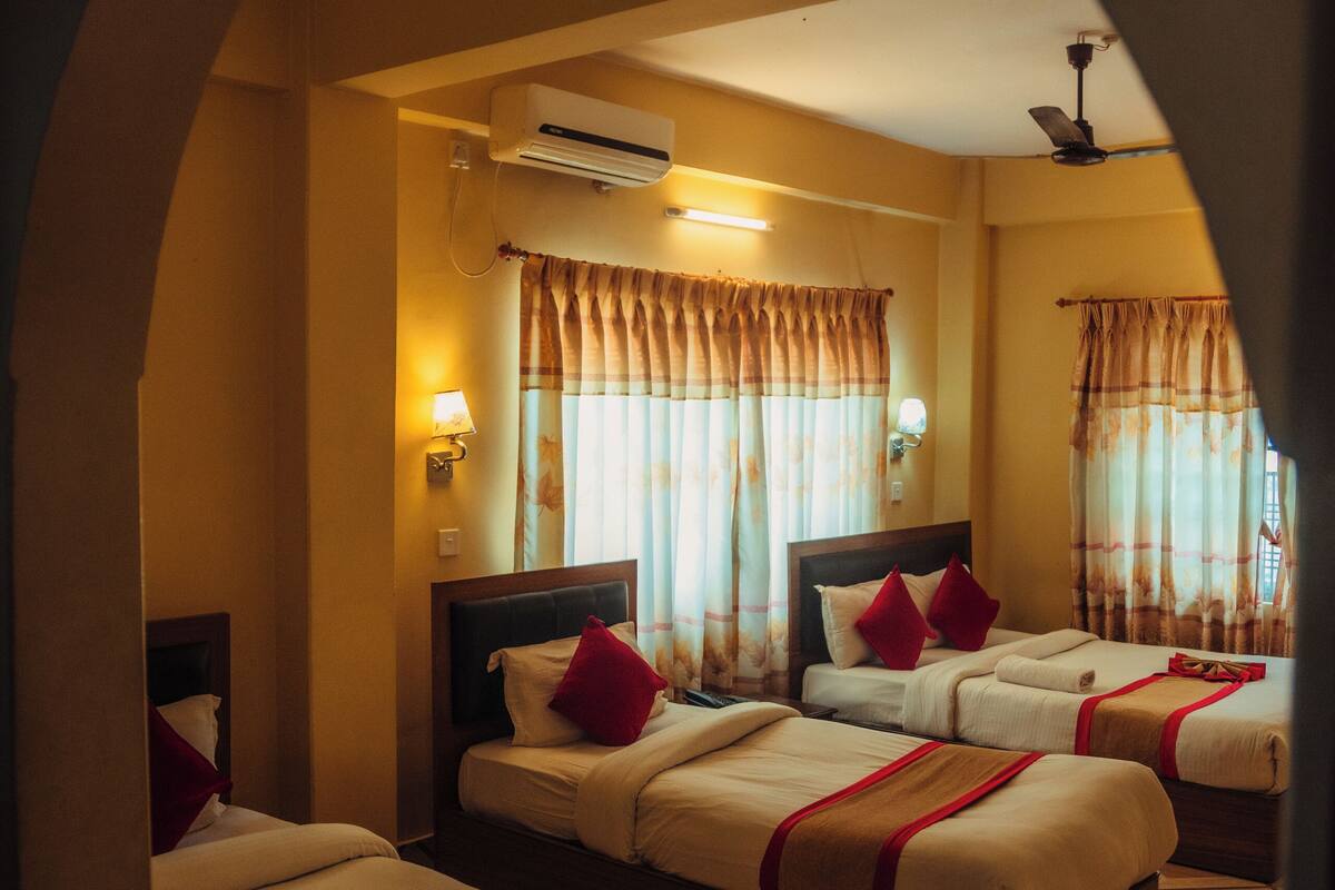 A hotel room with three beds, each with a red pillow and a decorative bow on the bedspread. The room has a tiled floor and a ceiling fan.