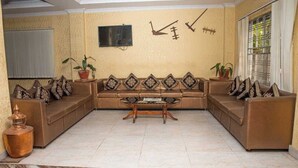 A living room with a brown sectional sofa, a wooden coffee table, and decorative pillows. There is a television mounted on the wall and a window with metal bars. - Hotel Gantabya Inn (Pokhara)