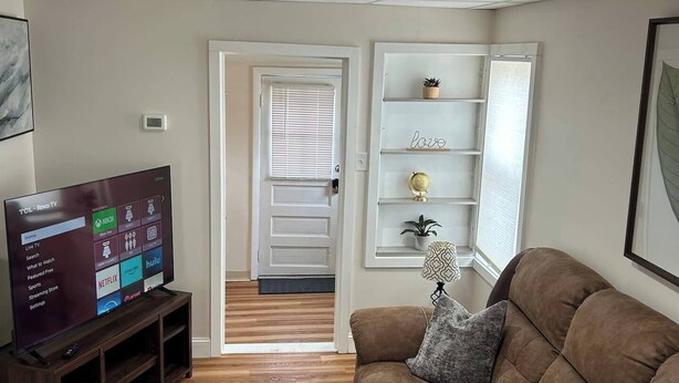 A living room with a brown sofa, a flat-screen TV on a stand, a patterned rug, and a shelving unit with decorative items.
