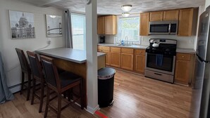 A kitchen with wooden cabinets, a stainless steel refrigerator, a microwave, and a stove. There are framed pictures on the wall, a window with blinds, and a trash can.