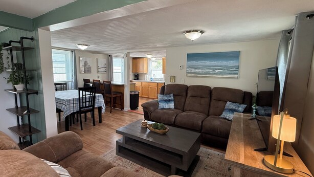 A living room with a brown sofa, a coffee table, and a lamp. There is a dining area with a table and chairs, and a kitchen in the background.