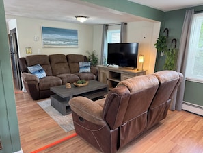Ein Wohnzimmer mit braunem Sofa, einem Couchtisch, einem Flachbildfernseher und einem Gemälde vom Strand.