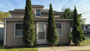 A two-story house with a garage door, a small porch, and four tall evergreen trees.