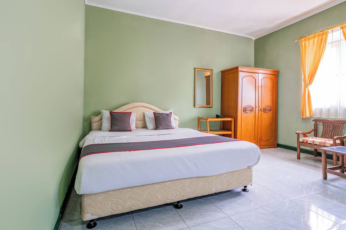 A neatly made bed with a headboard, a wooden wardrobe, and a chair in a room with green walls and tiled flooring.
