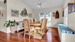 A dining area with a wooden table, chairs, and a bench. There are wall decorations, a ceiling fan, and a window with blinds. - Seaing is Believing | Surf City, NC | 4 Bedrooms | Short Walk to Beach (Surf City)