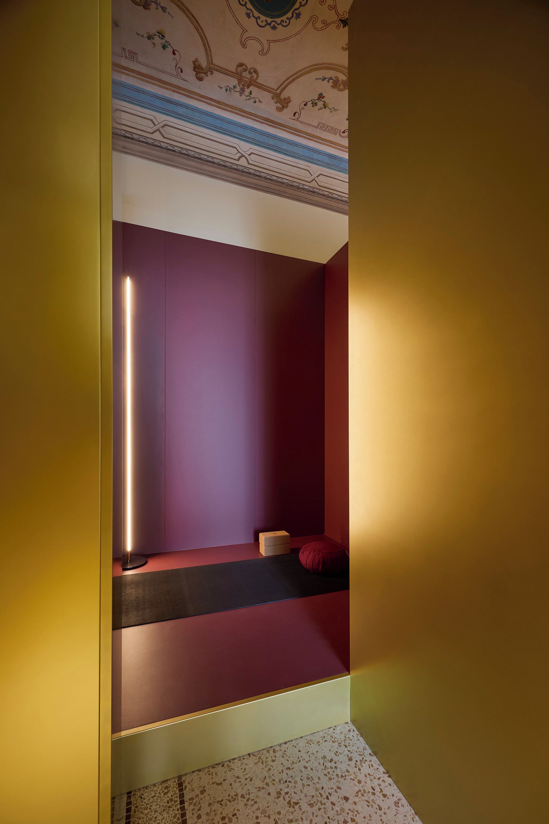 Une pièce au design moderne, avec un mur violet, un petit coin salon comprenant un banc et des coussins, et un plafond décoratif orné de motifs complexes.