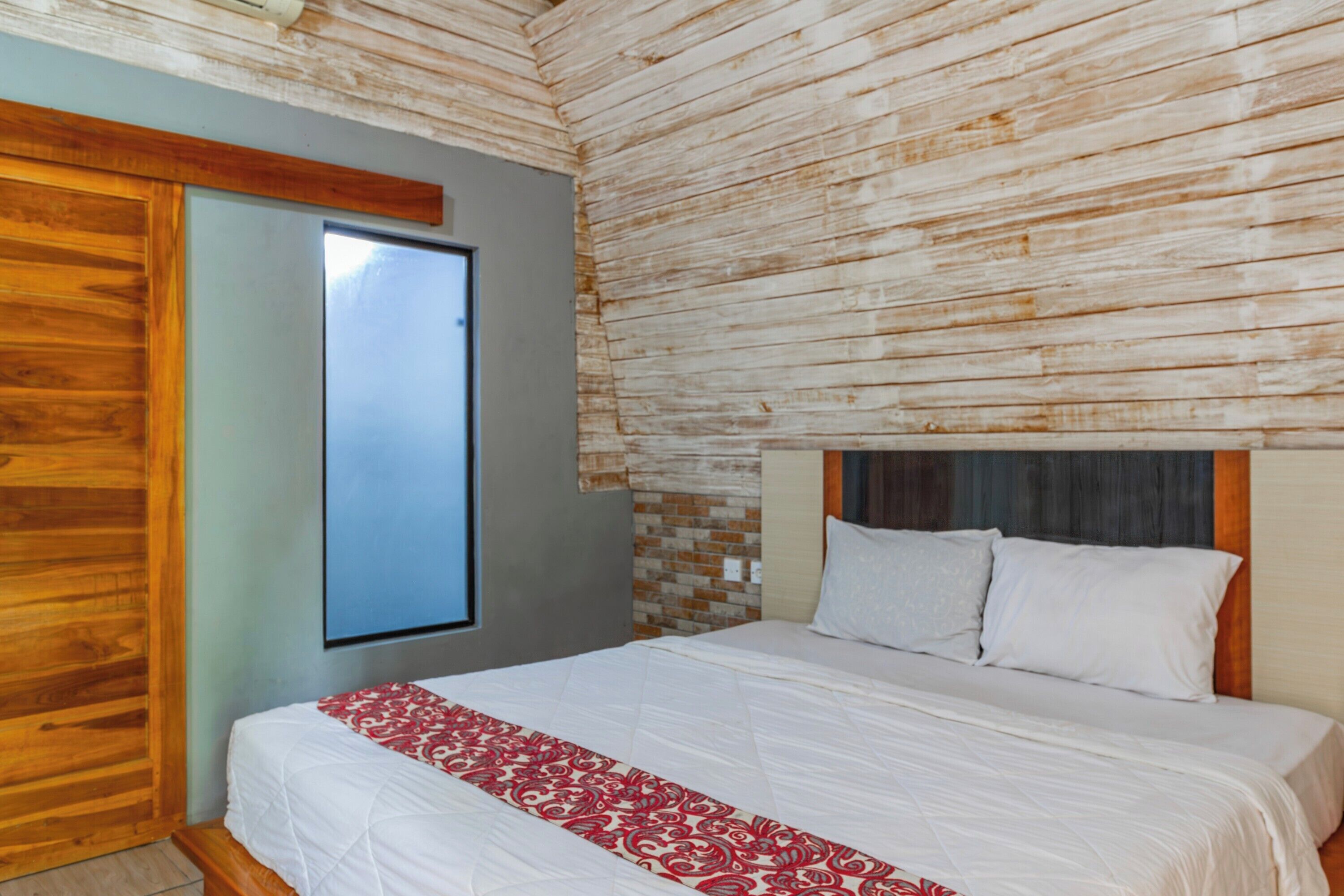 Un dormitorio con cabecera de madera, un ventanal grande y una cama con ropa de cama blanca y una manta roja y blanca estampada.
