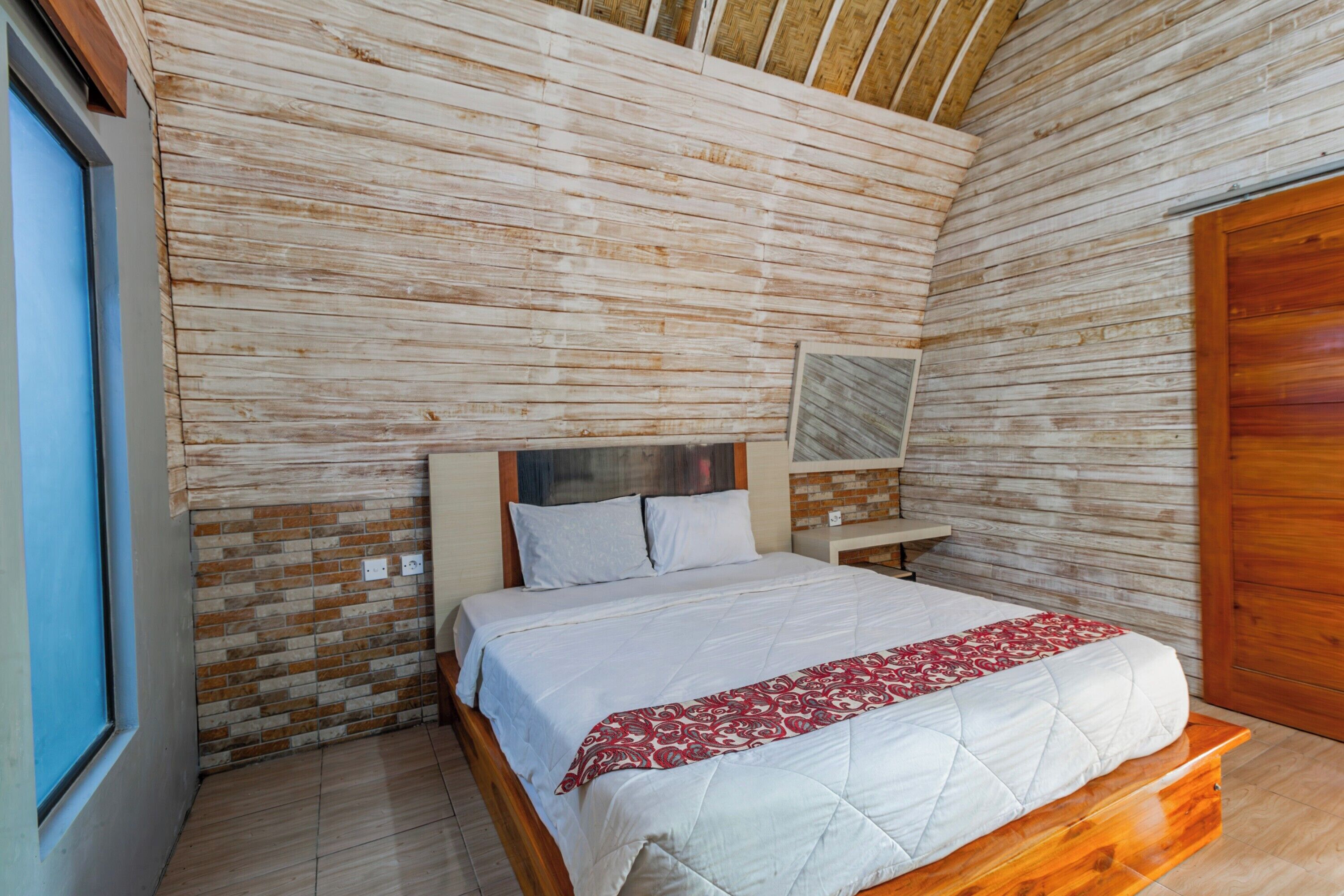 Une chambre avec un plafond en lambris de bois, un lit recouvert d’une courtepointe blanche matelassée et d’un plaid à motifs rouges, une table de chevet en bois et un mur en briques.