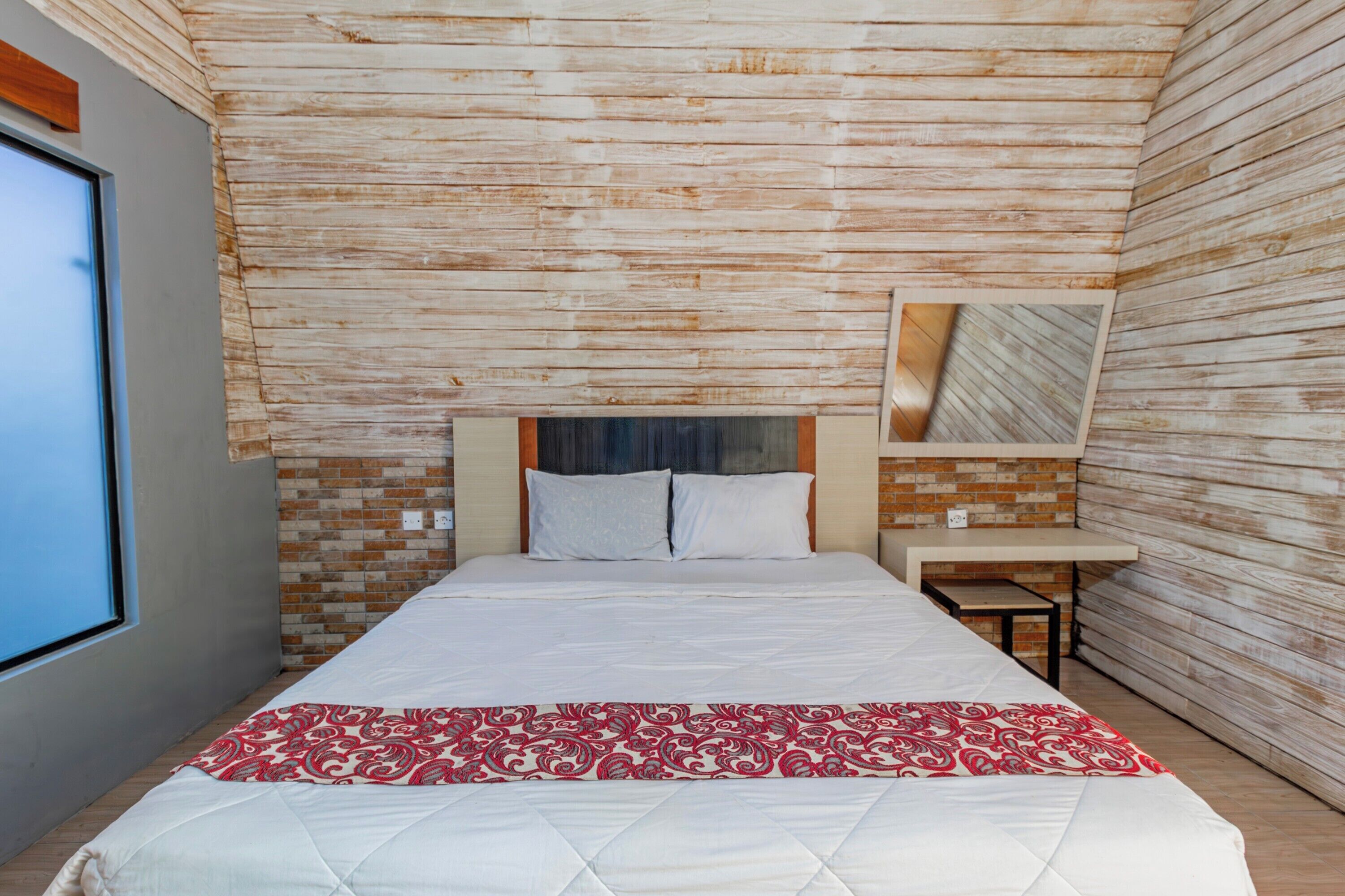 Un dormitorio moderno con techo de ladrillo, una cama grande con colcha blanca y roja, una mesita de noche de madera y un ventanal amplio.