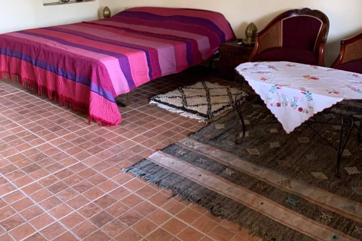 Une chambre à coucher avec un lit, une chaise, une table recouverte d’une nappe, un tapis, un tableau accroché au mur et un miroir.