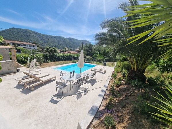 Un espace piscine avec des chaises longues et une table, entouré de verdure et avec une montagne en arrière-plan.