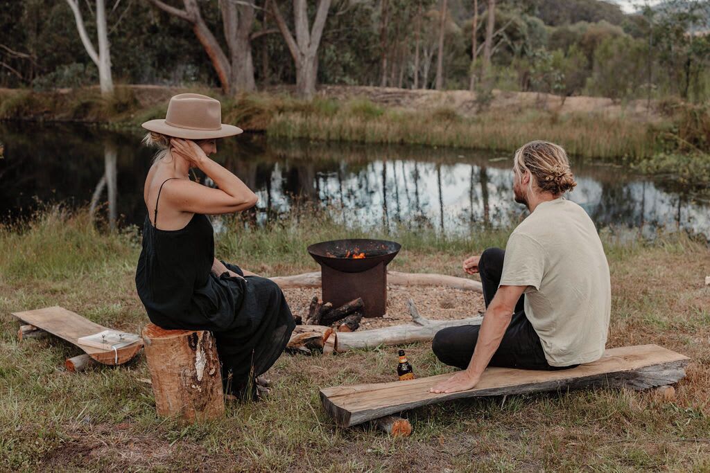Deux personnes sont assises près d’un feu de camp, dans un cadre naturel, avec un étang en arrière-plan.