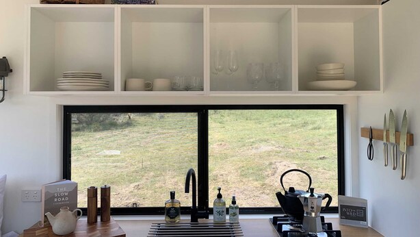 A modern kitchen with a large window, white cabinetry, and a variety of kitchenware. - Tiny Aura. (Coxs Crown)