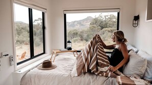 A bedroom with a large window, a bed, a bedside table with a lamp, and a view of the outdoors. - Tiny Aura. (Coxs Crown)